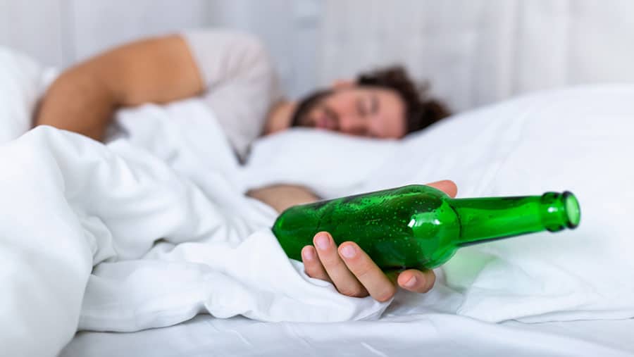 A man who is experiencing black out drinking is asleep in bed with a green alcohol bottle in hand.