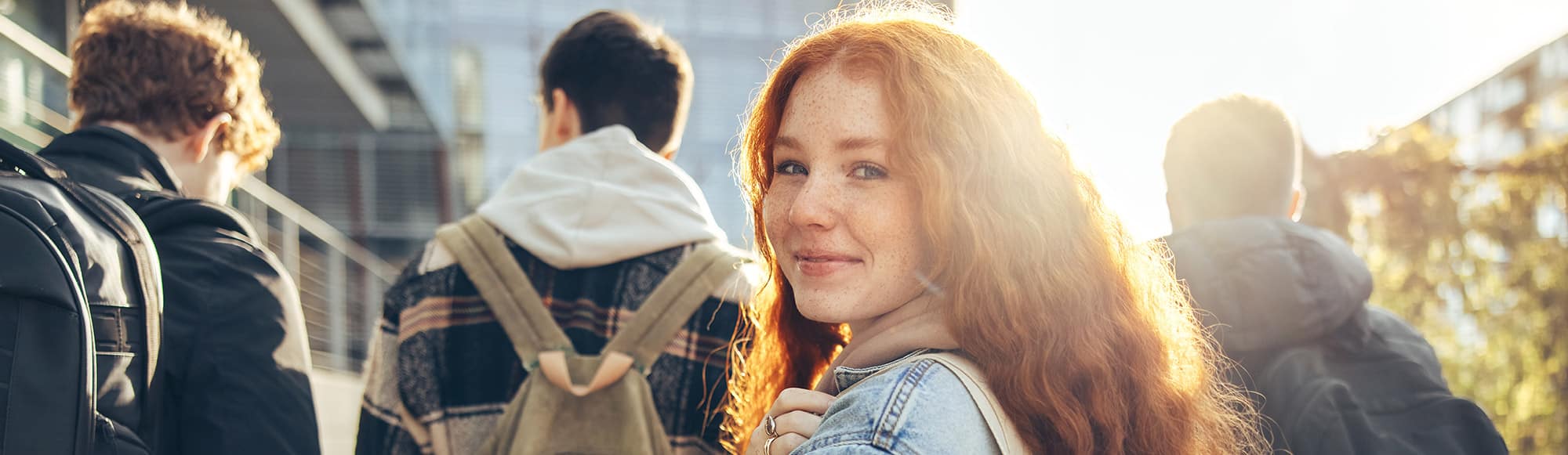 Younger, red haired girl walking behind younger adults casually dressed, looking back straight with the sun shining on her hair