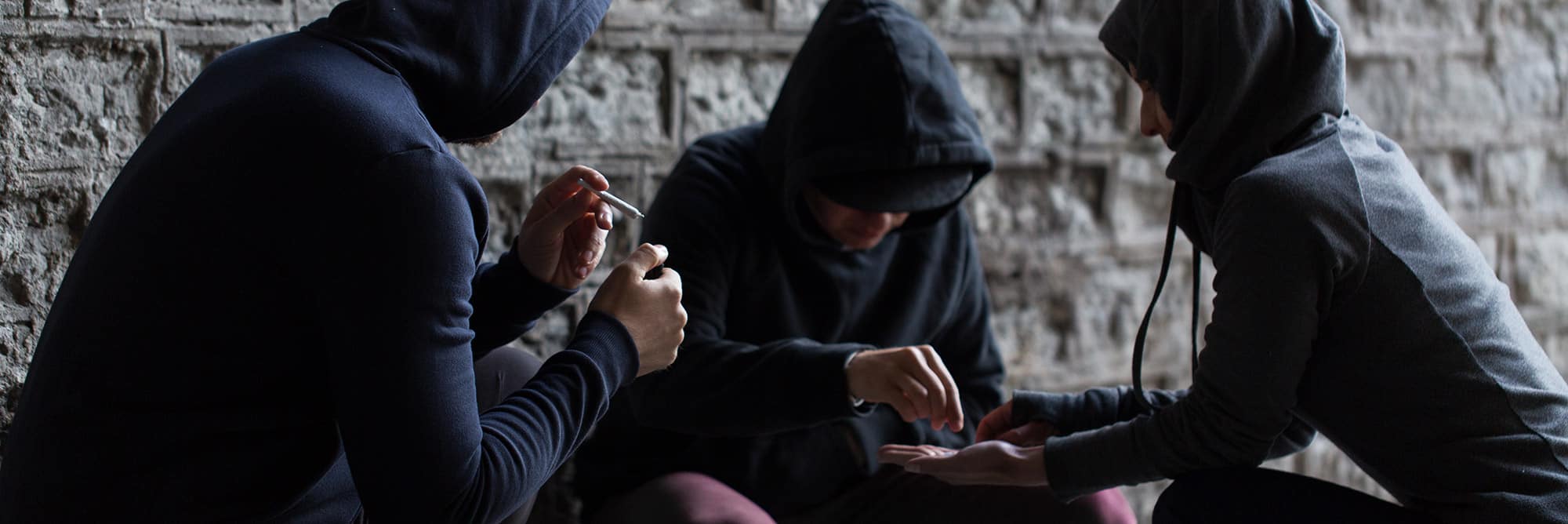 three young adults in need of rehab, huddles together near a dark brick wall, all of them are wearing hoodies. One is smoking a marijuana joint, the other is selling pills to the other