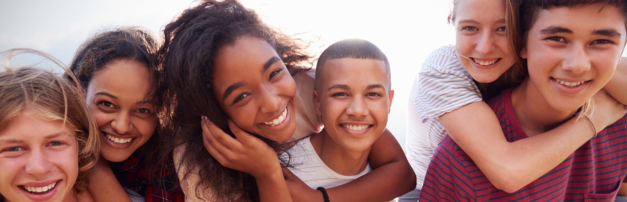 Close up of 6 casually dressed young adults, smiling and hugging, outside with the sunshine behind them