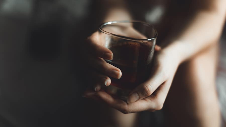 In a dark room a person is holding a glass cup of alcohol while contemplating what meds for alcohol withdrawal will be safe. 