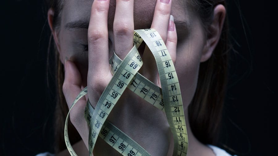 A person holding their hand to their face and entangled in their hand is a seamstress measuring tape representing Eating Disorder Awareness Week.