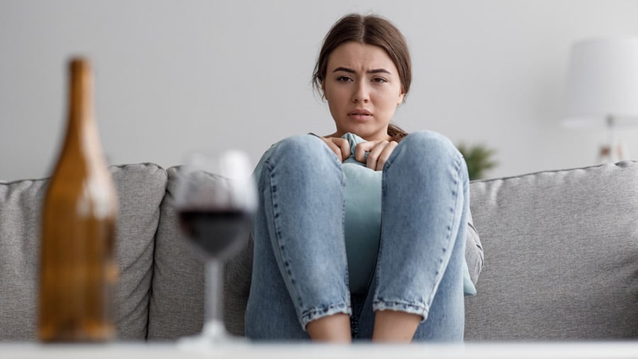 A young woman staring at a glass and bottle of wine in front of her represents the ways how to help an alcoholic friend recover from addiction.