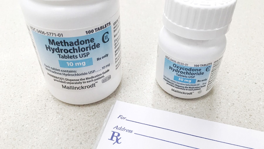 A white and blue medication bottle of methadone hydrochloride and a smaller bottle of oxycodone sit on a table. 