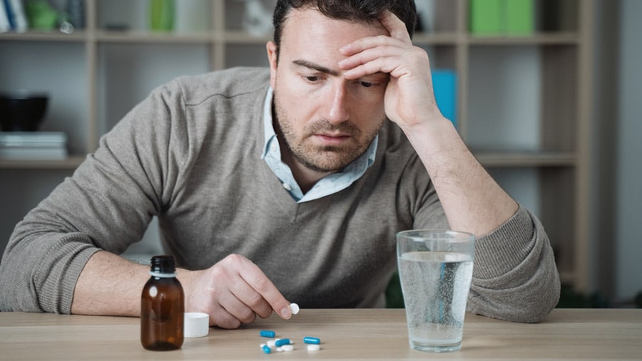 A man in a grey sweater sits at the table with blue and white pills in front of him. He contemplates his benzodiazepines addiction treatment. 