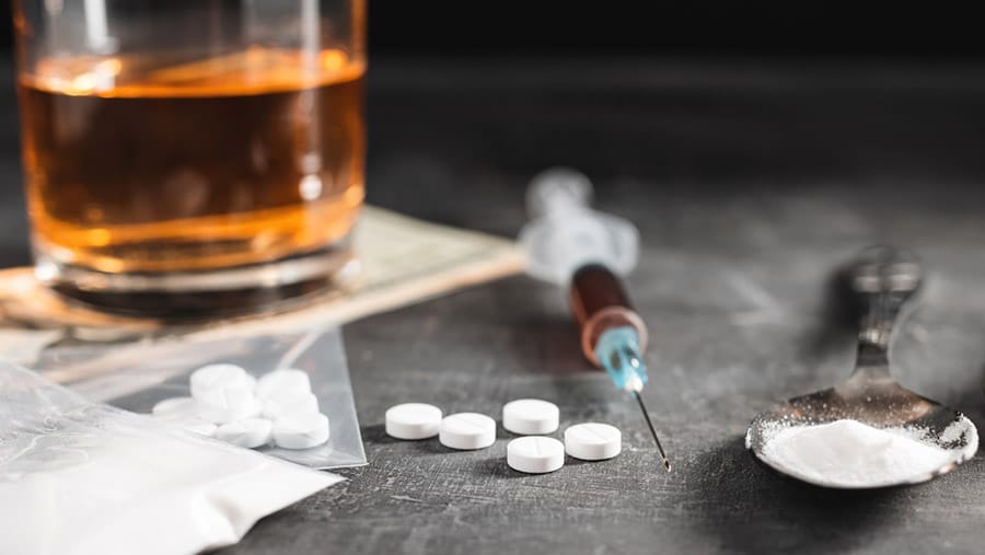 A blurred out glass of alcohol, a bag of white tablets, a syringe and a spoon filled with white powder represent polysubstance abuse of alcohol and heroin.