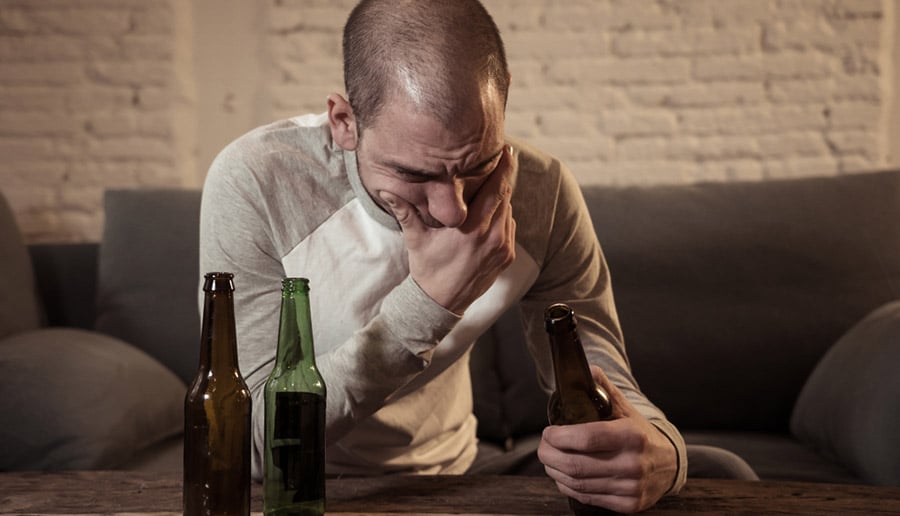 A man with a receding hair line holding a bottle of beer in his hands and contemplating if excessive drinking causes hair loss.