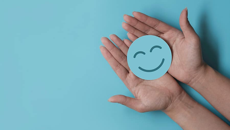 On a turquoise background, a person's hands holding a blue smiling face paper representing mental health misconceptions. 