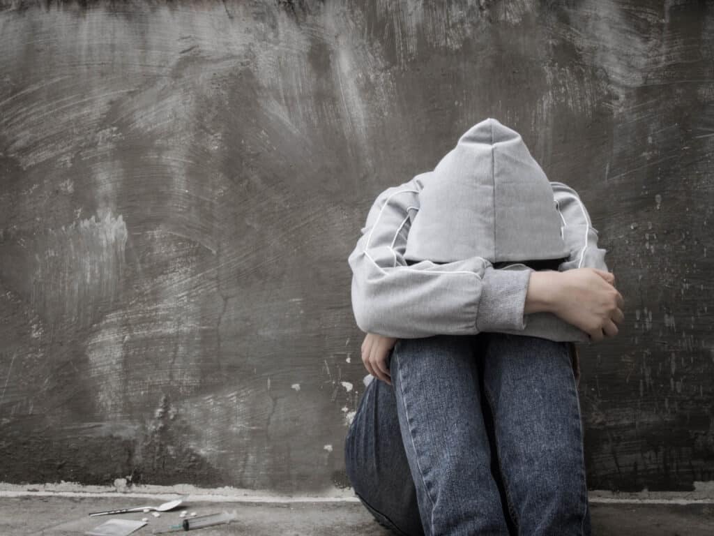 A man in grey hoodie with head in his arms sitting on the ground next to heroin pills, a spoon and syringe represents how to help a heroin addict.