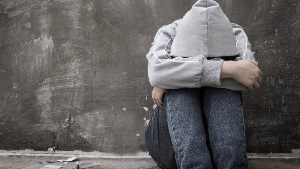 A man in grey hoodie with head in his arms sitting on the ground next to heroin pills, a spoon and syringe represents how to help a heroin addict.