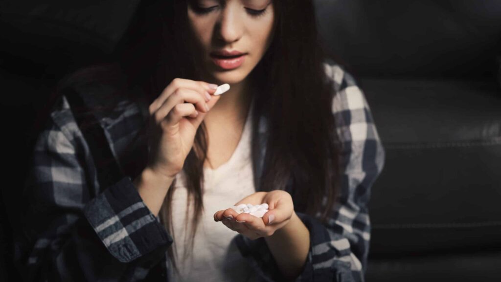 Young female in plaid shirt raising her hand to take a white klonopin pill while in the other hand holding the remaining white pills represents her klonopin addiction.