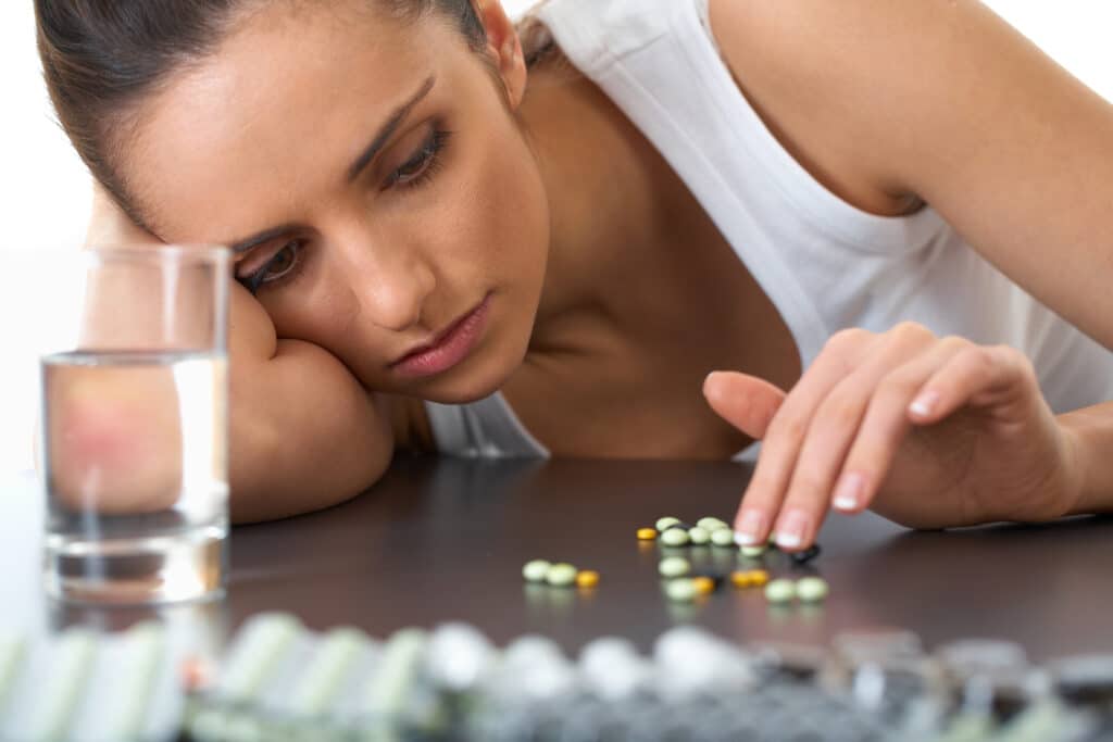 A young women suffering from substance use disorder and depression with her head on the table counting pills next to a glass of water.