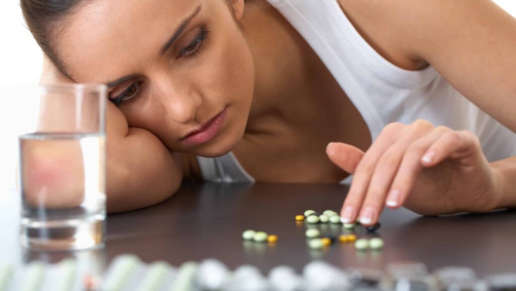 A young women suffering from substance use disorder and depression with her head on the table counting pills next to a glass of water.
