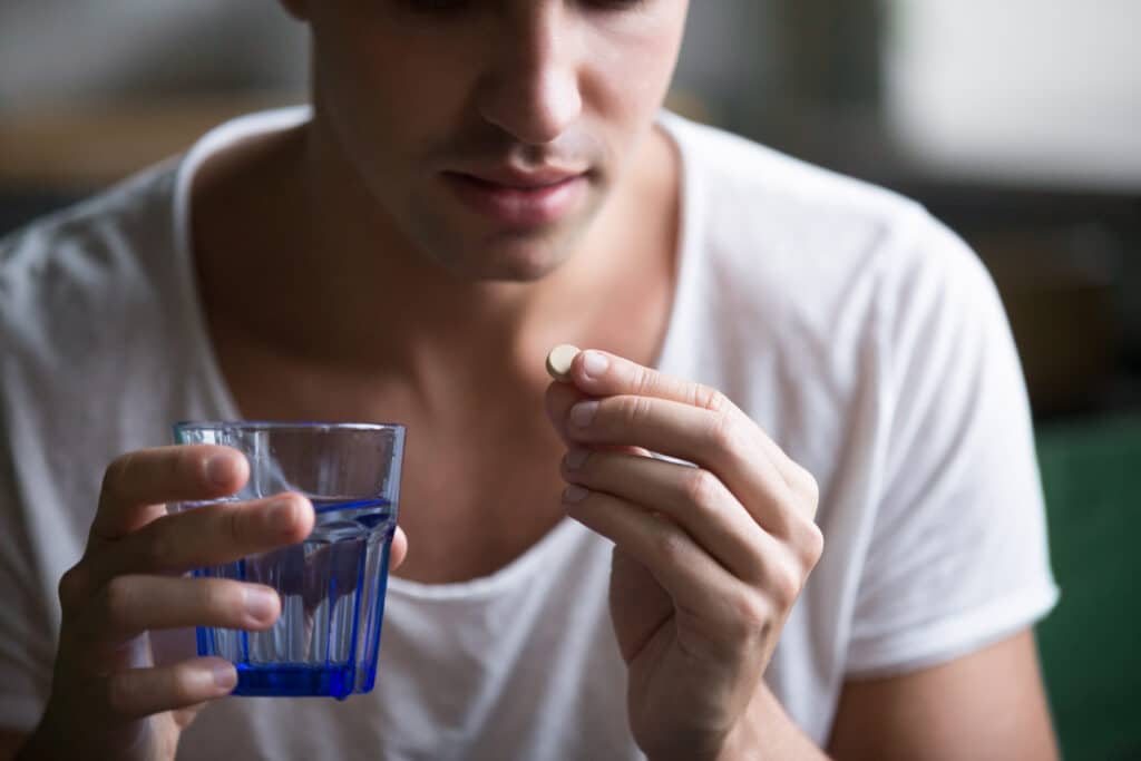 A young millennial man in a white shirt holding a glass of water and a suboxone tablet in the other hand is thinking about what medications can you not take with suboxone.