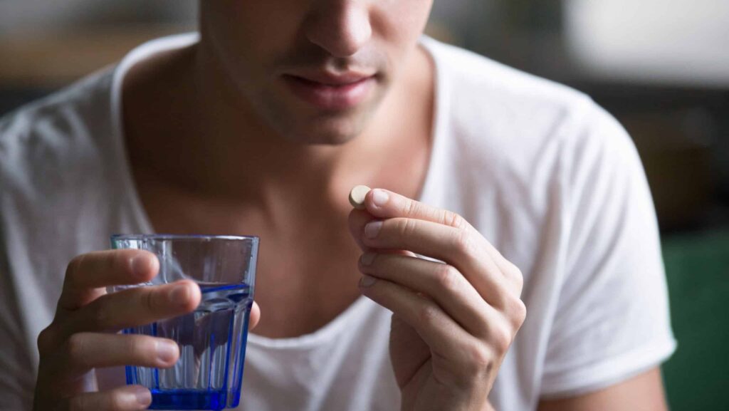 A young millennial man in a white shirt holding a glass of water and a suboxone tablet in the other hand is thinking about what medications can you not take with suboxone.