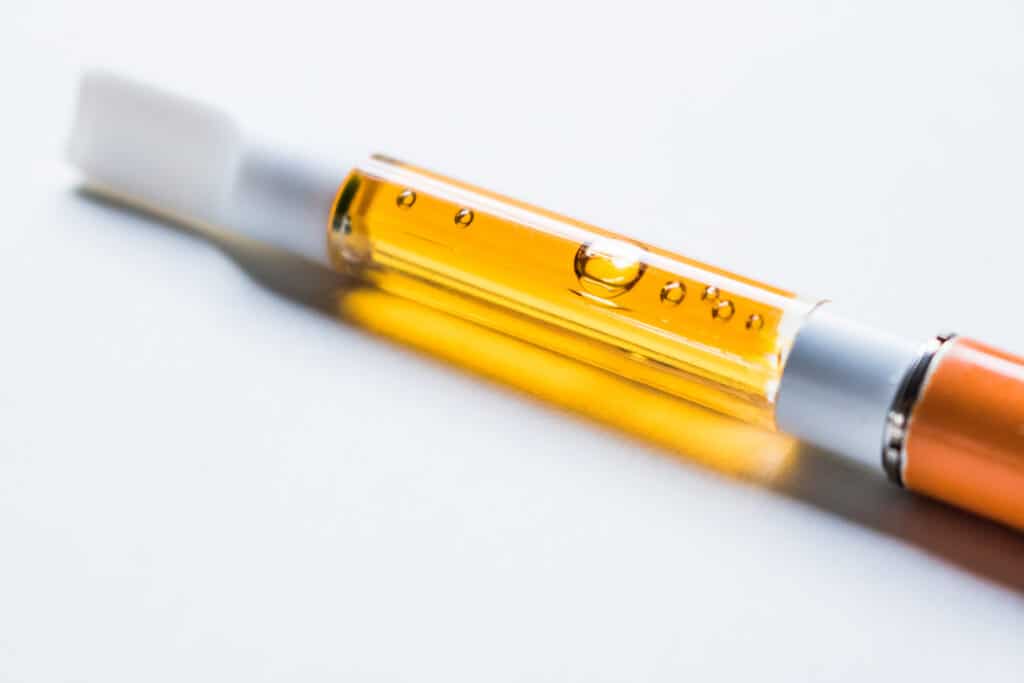 A white and orange disposable wax pen on a white background contains honey colored liquid cannabis.