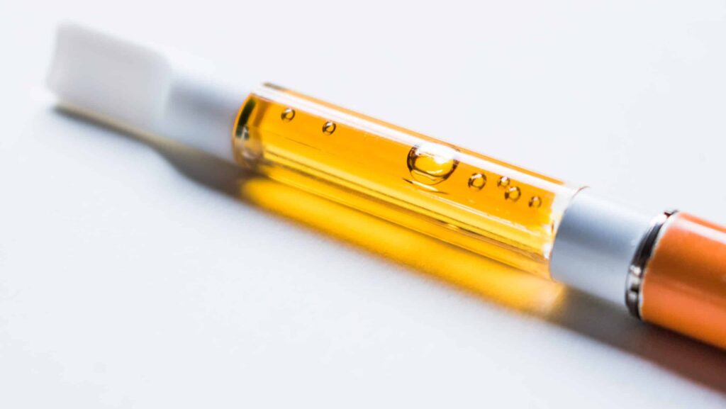 A white and orange disposable wax pen on a white background contains honey colored liquid cannabis.