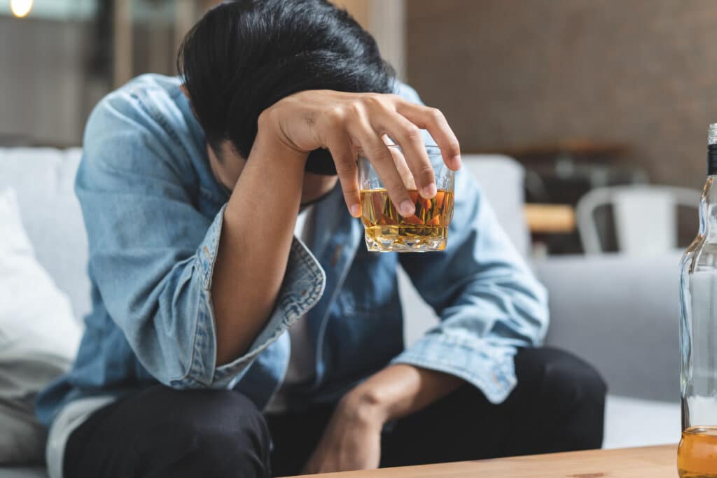 A young man in a denim shirt on a sofa with his head hung low holds a glass of alcohol and contemplates how to help an alcoholic who doesn't want help.