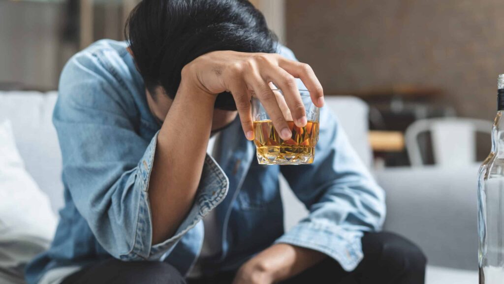 A young man in a denim shirt on a sofa with his head hung low holds a glass of alcohol and contemplates how to help an alcoholic who doesn't want help.