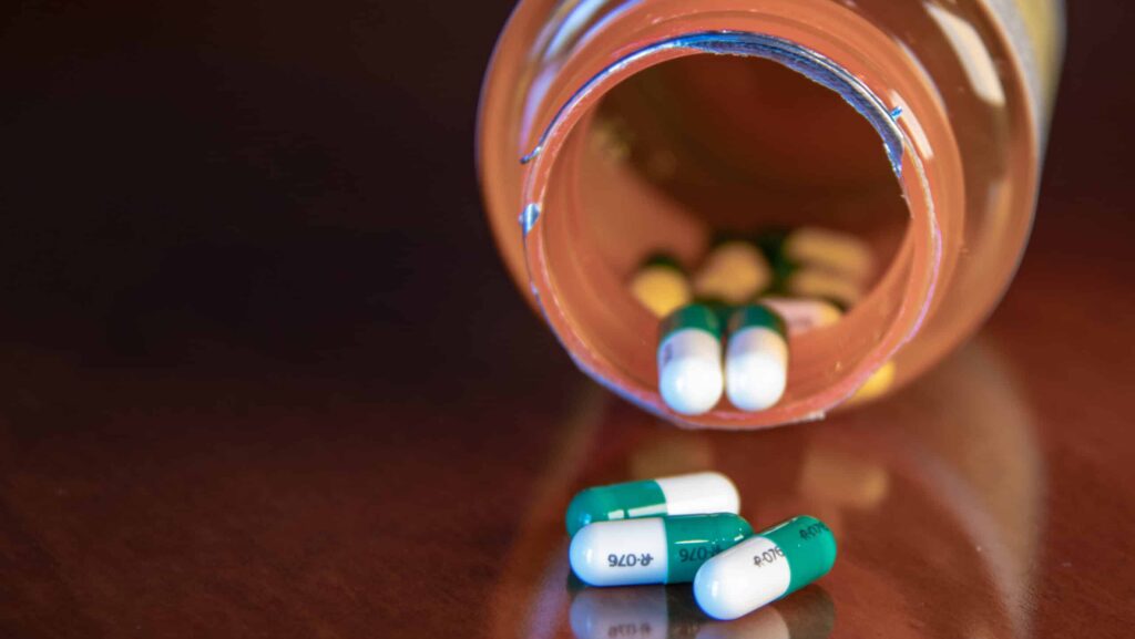 A tipped over pill bottle on a wooden table with green and white pills spilled out. It represents Restoril vs Ambien, both drugs to treat insomnia.