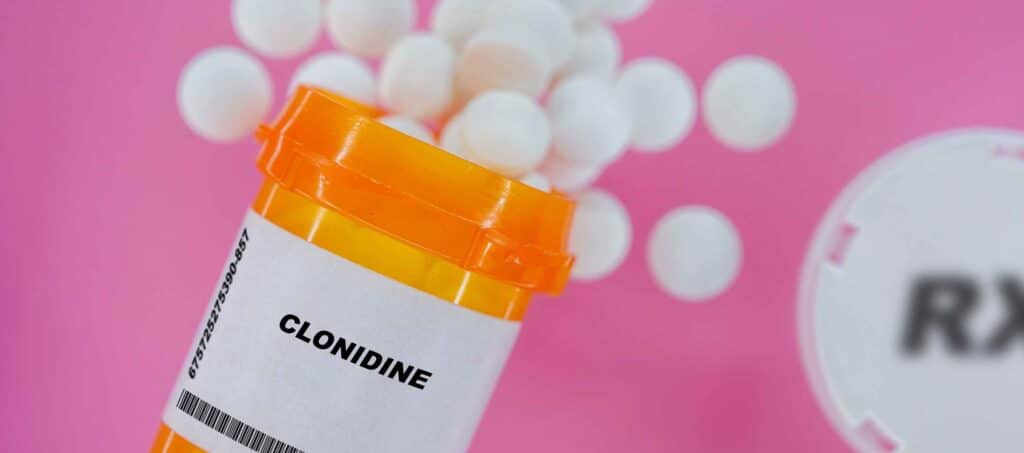 On a pink background, a pill bottle of Clonidine is spilling out white tablets representing Clonidine addiction.