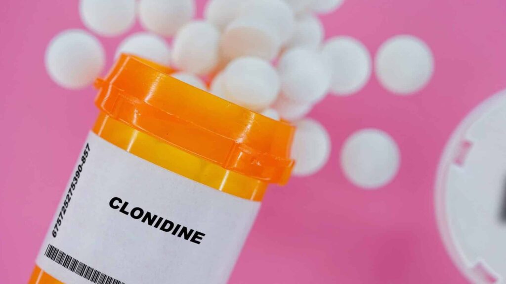 On a pink background, a pill bottle of Clonidine is spilling out white tablets representing Clonidine addiction.