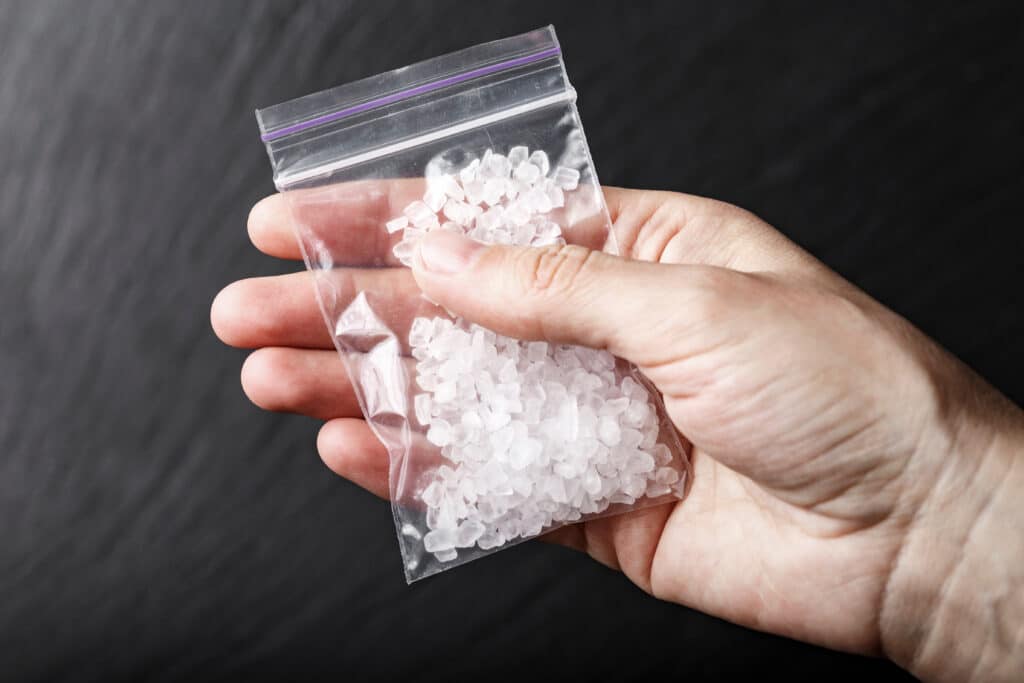 A hand is holding a small clear bag of crystal meth on a black background represents testing of how long does meth stay in your urine.