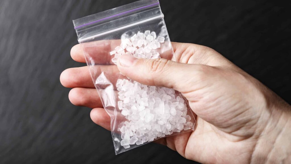 A hand is holding a small clear bag of crystal meth on a black background represents testing of how long does meth stay in your urine.