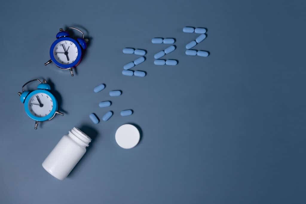On a blue background, there are two mini blue alarm clocks next to a white pill bottle spilling blue pills in the shape of a Z. The photo represents the question, can you overdose on sleeping pills?