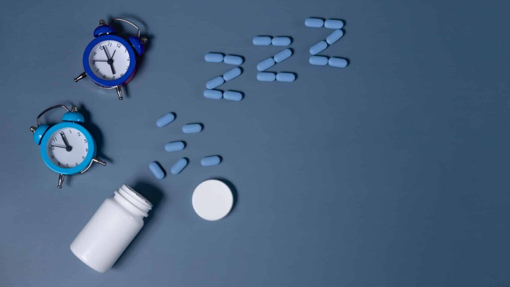 On a blue background, there are two mini blue alarm clocks next to a white pill bottle spilling blue pills in the shape of a Z. The photo represents the question, can you overdose on sleeping pills?