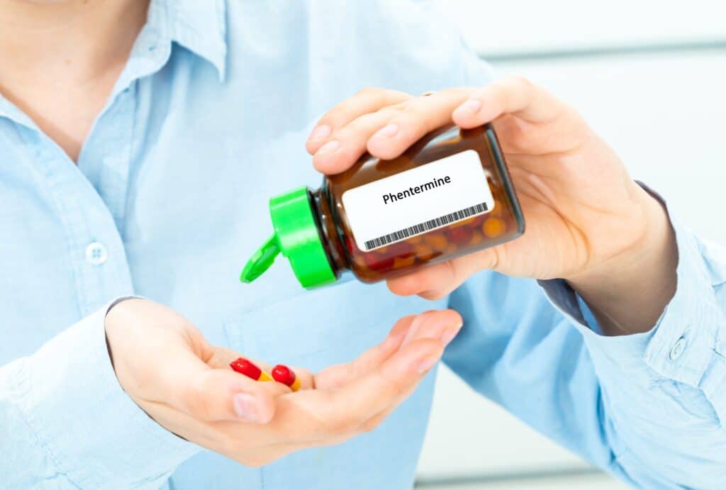 A person in a light blue shirt is holding a bottle of Phentermine and pouring out the pills into the other hand. This represents phentermine addiction.