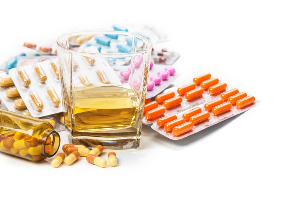 Multiple sleeping pills in varying colors are laid out next to a glass of alcohol representing the mixing of Ambien and alcohol.