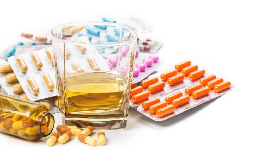 Multiple sleeping pills in varying colors are laid out next to a glass of alcohol representing the mixing of Ambien and alcohol.