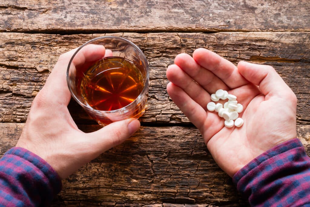 A man's hands on a wooden table is holding a glass of alcohol in one and pills in the other questions how long after drinking can you take xanax.