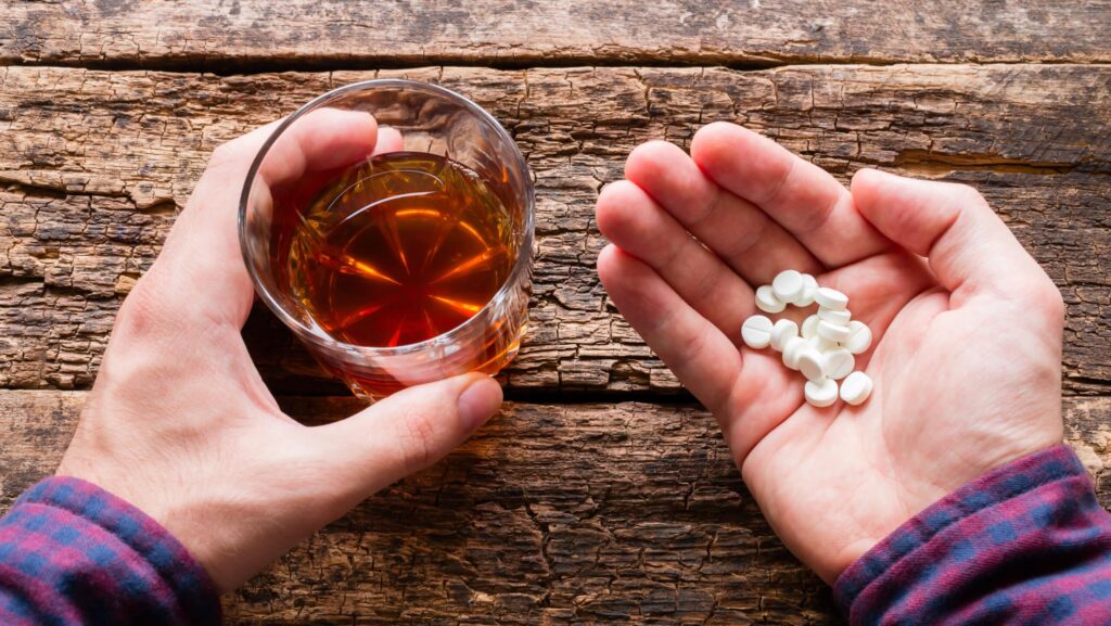 A man's hands on a wooden table is holding a glass of alcohol in one and pills in the other questions how long after drinking can you take xanax.