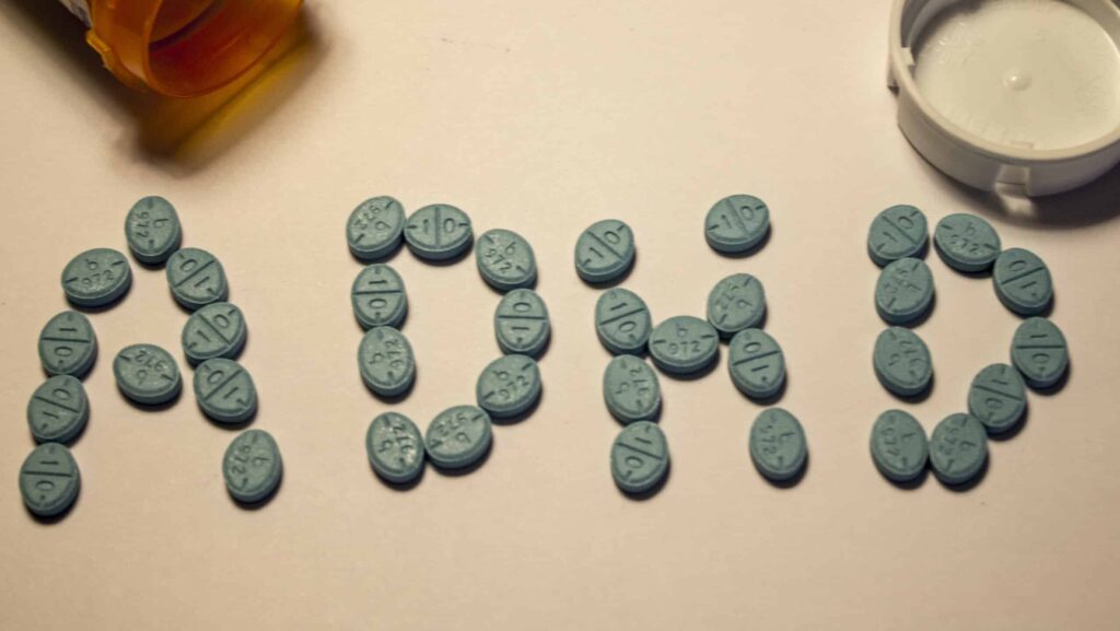 On a white table with a pharmacy pill bottle tipped over, blue Adderall pills are arranged in to the letters ADHD. It represents the long term effects of Adderall.