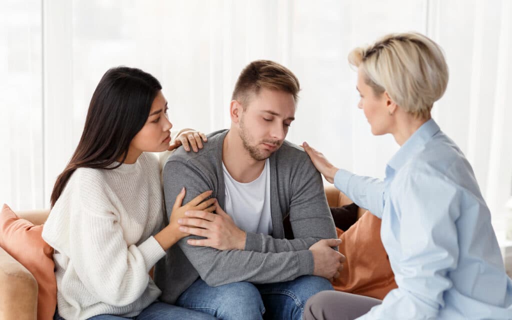 A young man with a drug addiction being consoled by two female friends represents the difference between enabling an addict and supporting an addict.