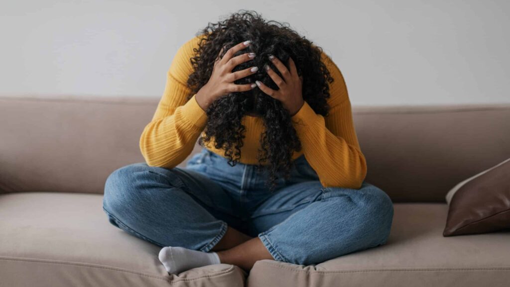 A young woman sitting on a sofa with her hands on her bent down head is an ideal candidate for ketamine treatment for PTSD.