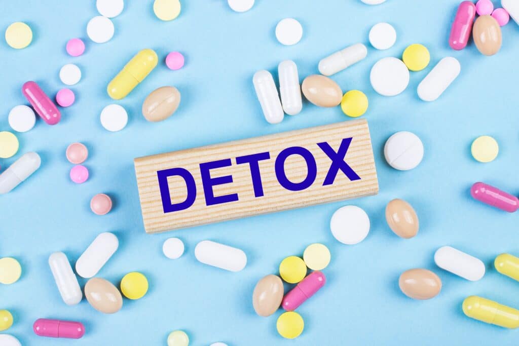 On a light blue background, the word detox printed on a wood block is surrounded by multicolored pills represents how to detox from meth.