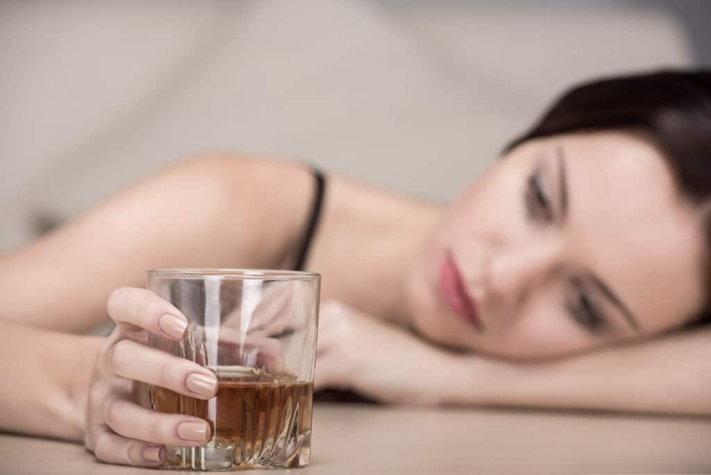 A young depressed female has her head on her arm while holding a glass of alcohol with the other hand on the table representing trazodone and alcohol use.