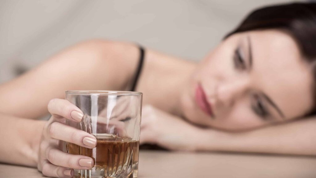 A young depressed female has her head on her arm while holding a glass of alcohol with the other hand on the table representing trazodone and alcohol use.