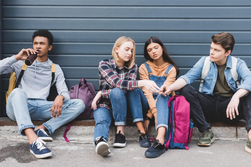 A group of young people sitting on a curb against a blue wall drinking and smoking answering the question of what is the number one drug used by teens.