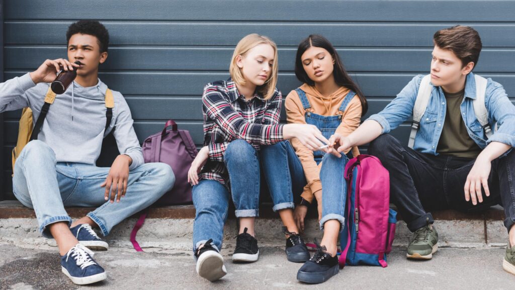 A group of young people sitting on a curb against a blue wall drinking and smoking answering the question of what is the number one drug used by teens.