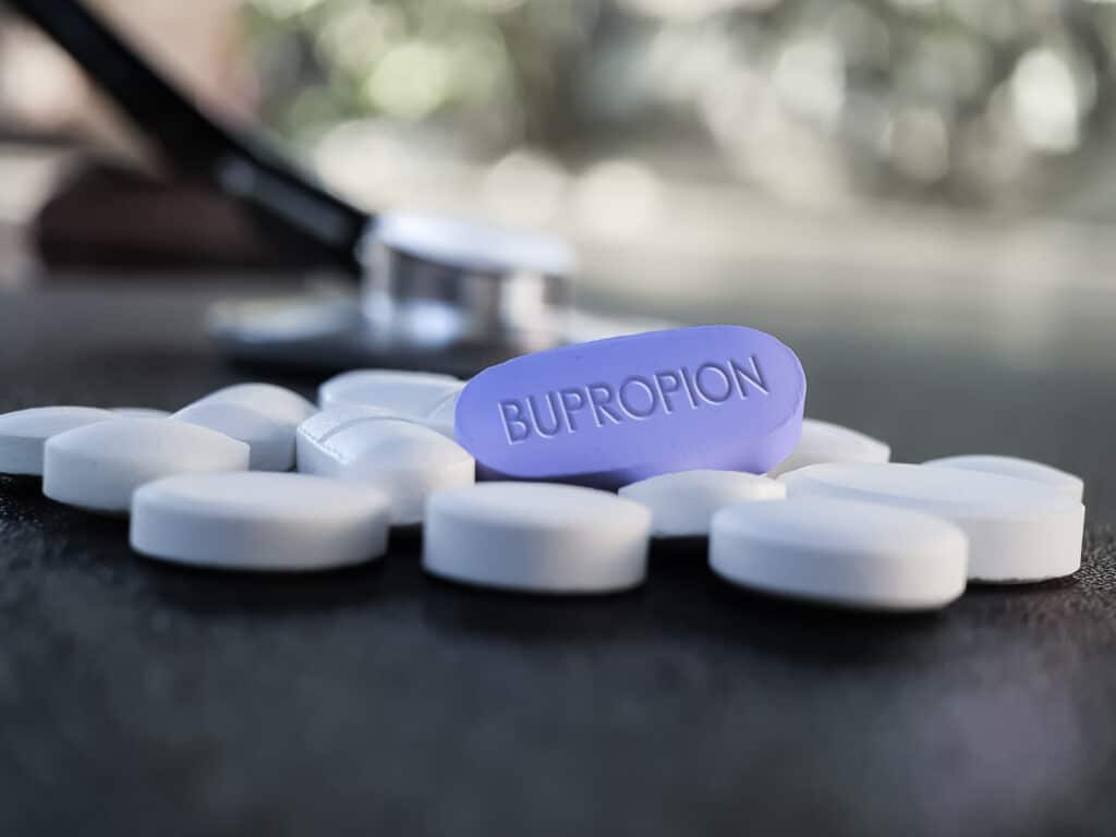 On a dark table, white tablets surround a purple tablet labeled bupropion represent what the bupropion withdrawal timeline might look like.