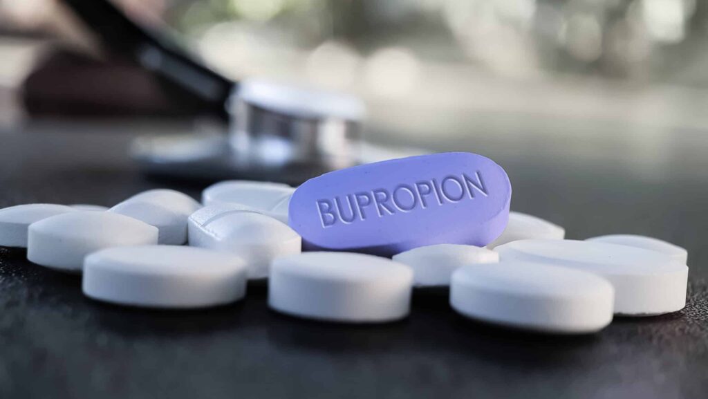 On a dark table, white tablets surround a purple tablet labeled bupropion represent what the bupropion withdrawal timeline might look like.