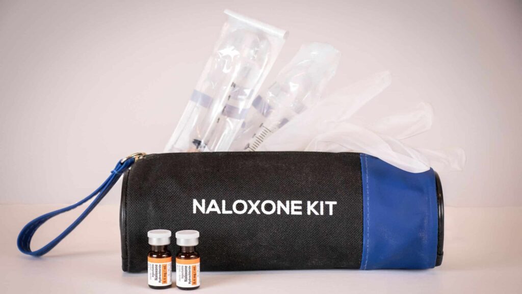 A black and blue bag labeled naloxone kit filled with clean syringes and gloves laying next to two bottles of naloxone represent what you may find at a harm reduction action center.