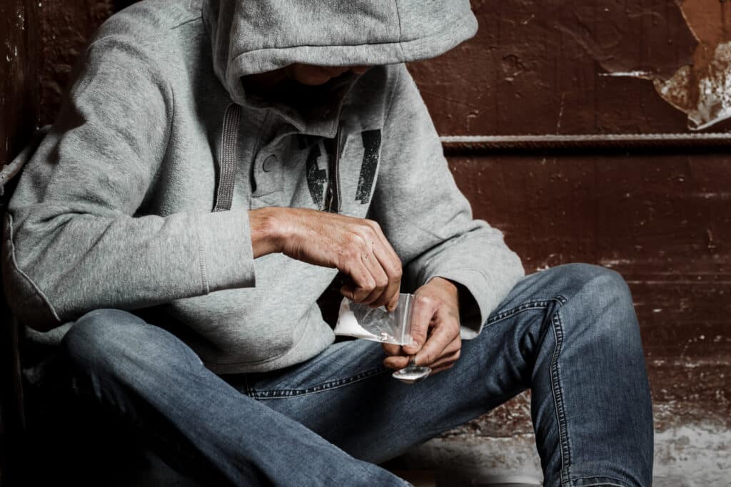 A man in a gray hoodie is seen pour white powdered heroin into a spoon representing the symptoms of heroin withdrawal.