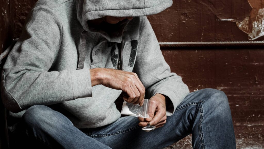 A man in a gray hoodie is seen pour white powdered heroin into a spoon representing the symptoms of heroin withdrawal.