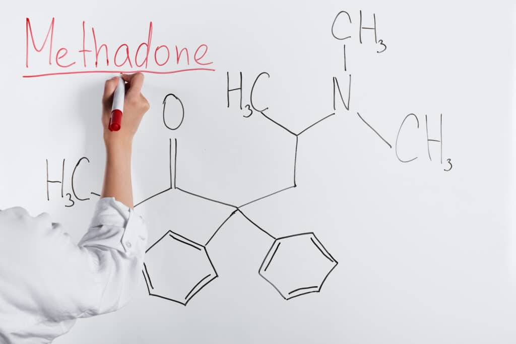 A woman in a white shirt writes the word methadone and draws its chemical structure on a white board to discuss methadone overdose symptoms.
