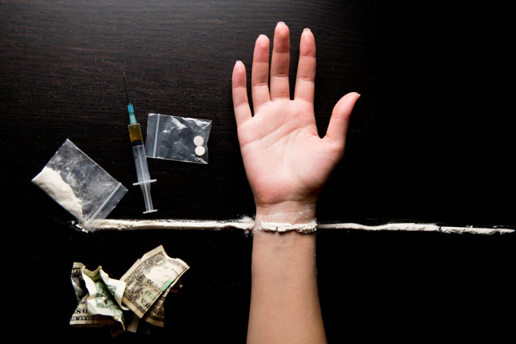 A persons hand is laid out on a dark table next to a syringe, cash, bags of drugs and powdered drugs, which represents meth vs crack similarities and differences.